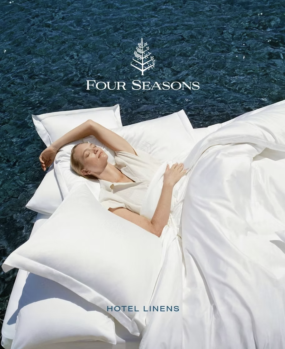 Woman relaxing in a white linen bed set over deep blue ocean water for Four Seasons Hotel Linens.