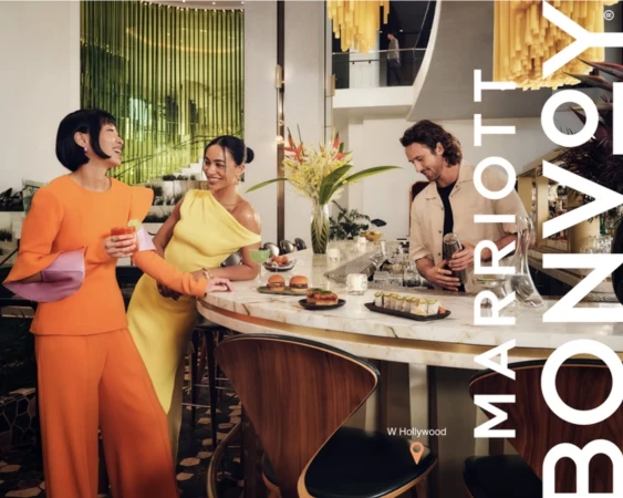 Two women in vibrant orange and yellow outfits conversing at a curved marble hotel bar while a bartender prepares drinks.