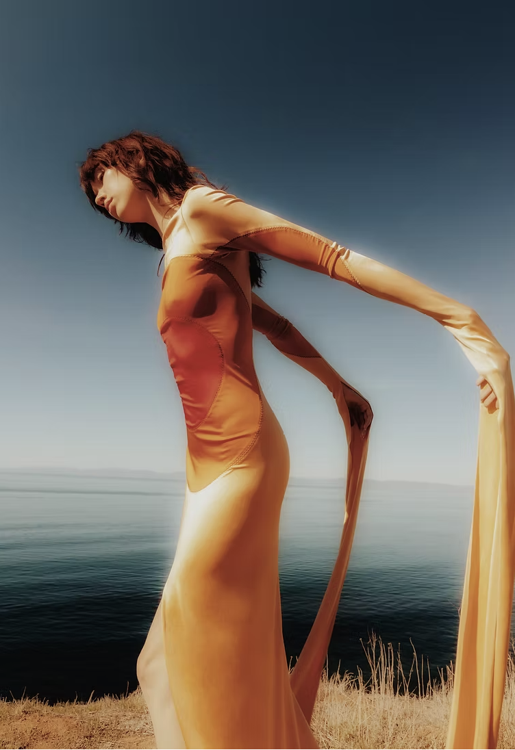 Model in an orange and yellow gradient dress with elongated flowing sleeves posing on a cliff above the ocean.
