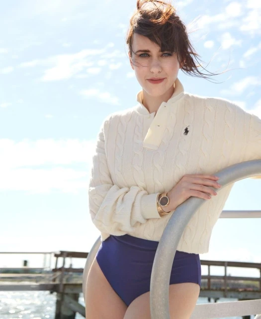 Woman in polo sweater and swimsuit bottoms poses on pier with ocean backdrop under bright sky and sunlight. Pier railing in foreground.