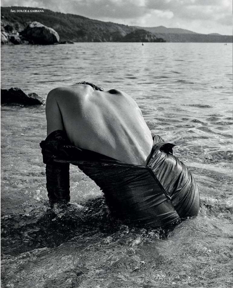 Monochrome image of a shirtless person with back to camera, wading in water, dark fabric draped low, ocean and island background.