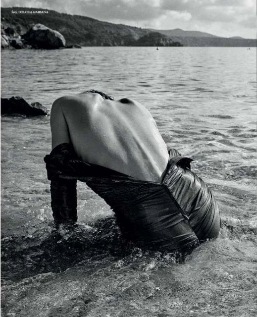 Monochrome image of a shirtless person with back to camera, wading in water, dark fabric draped low, ocean and island background.