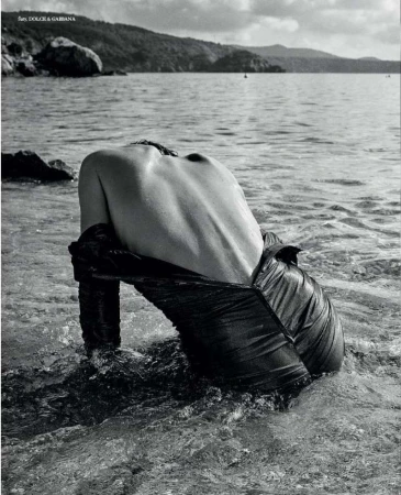 Monochrome image of a shirtless person with back to camera, wading in water, dark fabric draped low, ocean and island background.