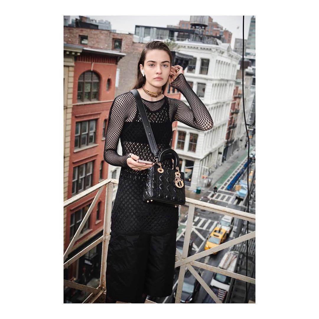 Fashion shot of a woman in a black fishnet top and skirt with a handbag on a city balcony overlooking a street and buildings.