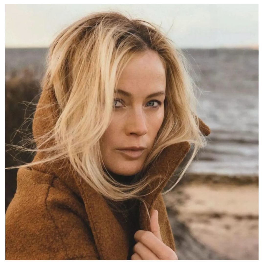 Portrait of a blonde woman in a brown hooded coat standing near the ocean with wavy hair and a neutral expression in natural light.