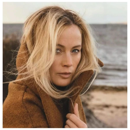 Portrait of a blonde woman in a brown hooded coat standing near the ocean with wavy hair and a neutral expression in natural light.