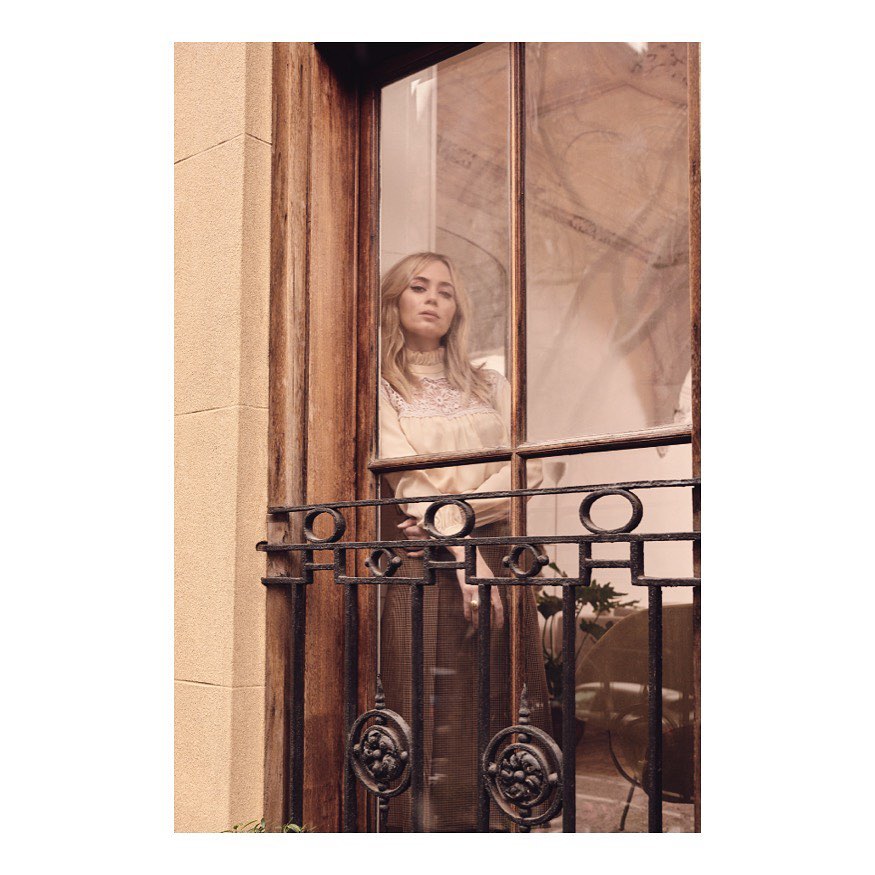 Adult woman with blonde hair stands on a balcony with wrought iron railing, posing in a creamy white long sleeve top.