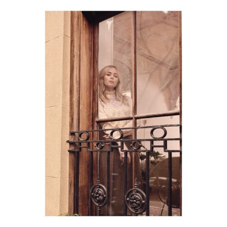 Adult woman with blonde hair stands on a balcony with wrought iron railing, posing in a creamy white long sleeve top.