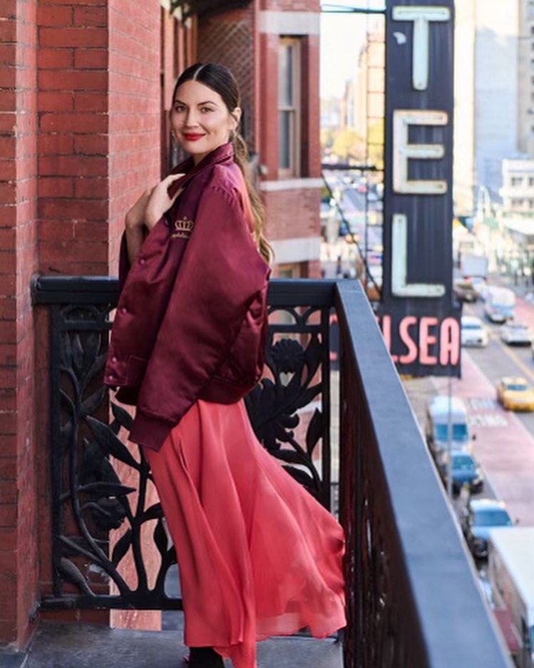 Adult woman poses on a balcony in a red dress and bomber jacket; city street and hotel signage visible in the background.