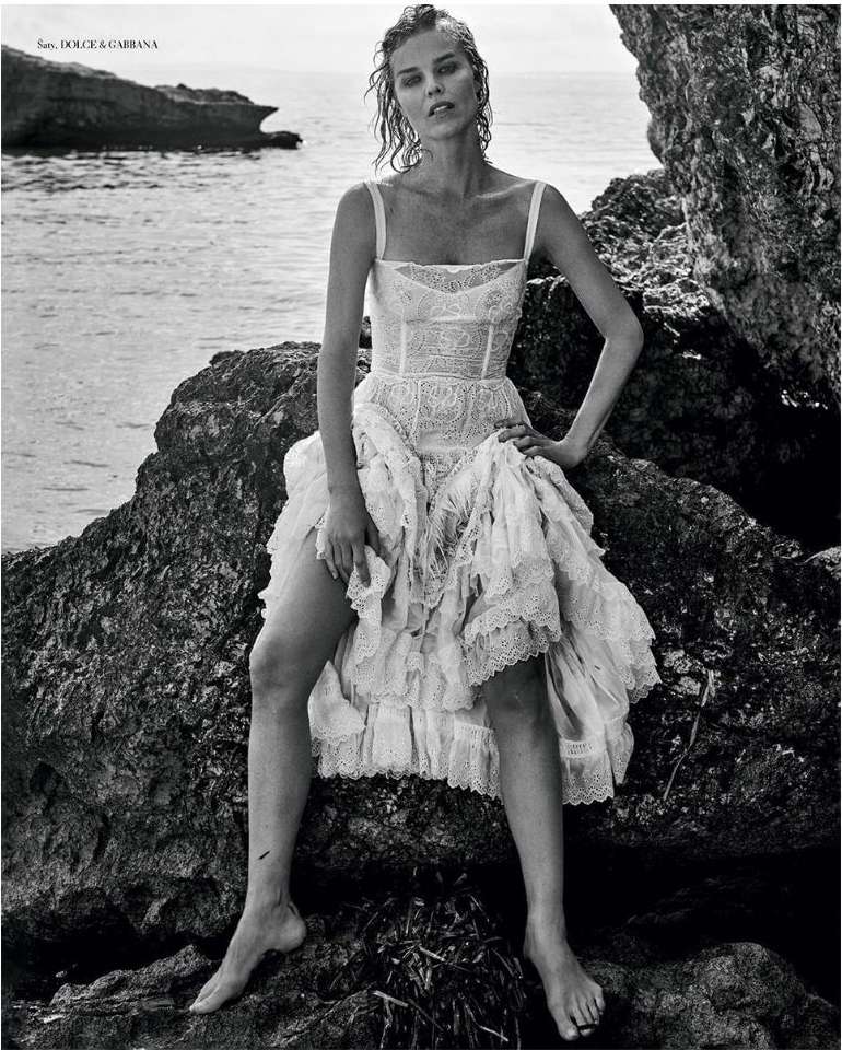 Monochrome fashion shot: a model in a white lace dress poses on rocks by the sea, creating a timeless and elegant portrait.
