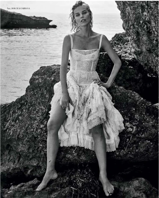 Monochrome fashion shot: a model in a white lace dress poses on rocks by the sea, creating a timeless and elegant portrait.
