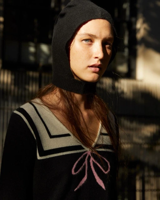A young adult female model with pale skin and freckles poses in a black balaclava and sweater with a pink bow, captured in natural light.