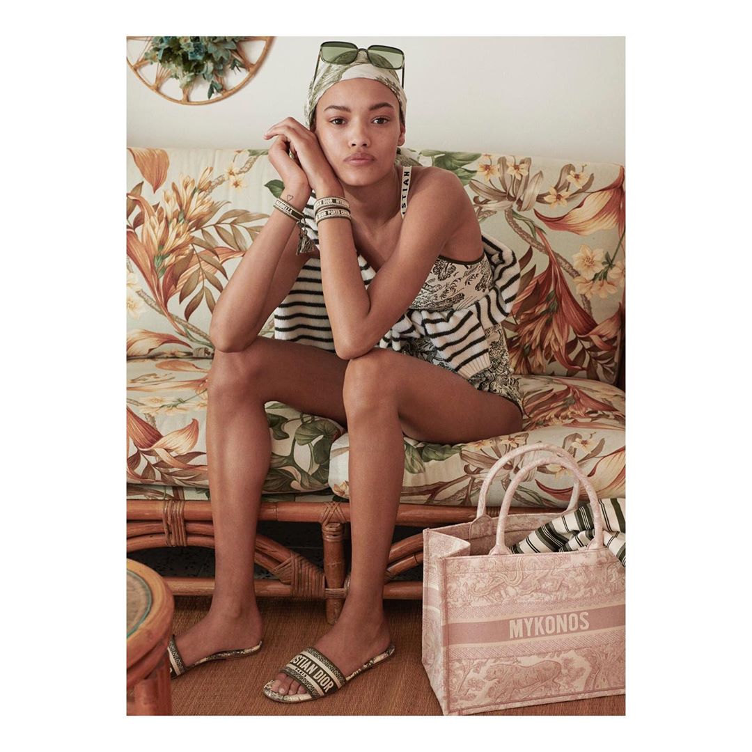 Fashion shot of a woman on a floral couch wearing a headscarf and slides, accessorized with bracelets, and a tote bag beside her.