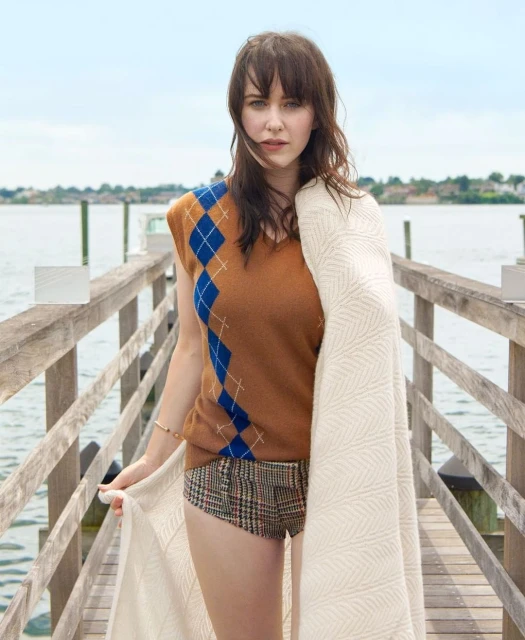 A young woman in a brown argyle vest and patterned shorts poses on a wooden pier, holding a white blanket with a serene waterfront view.