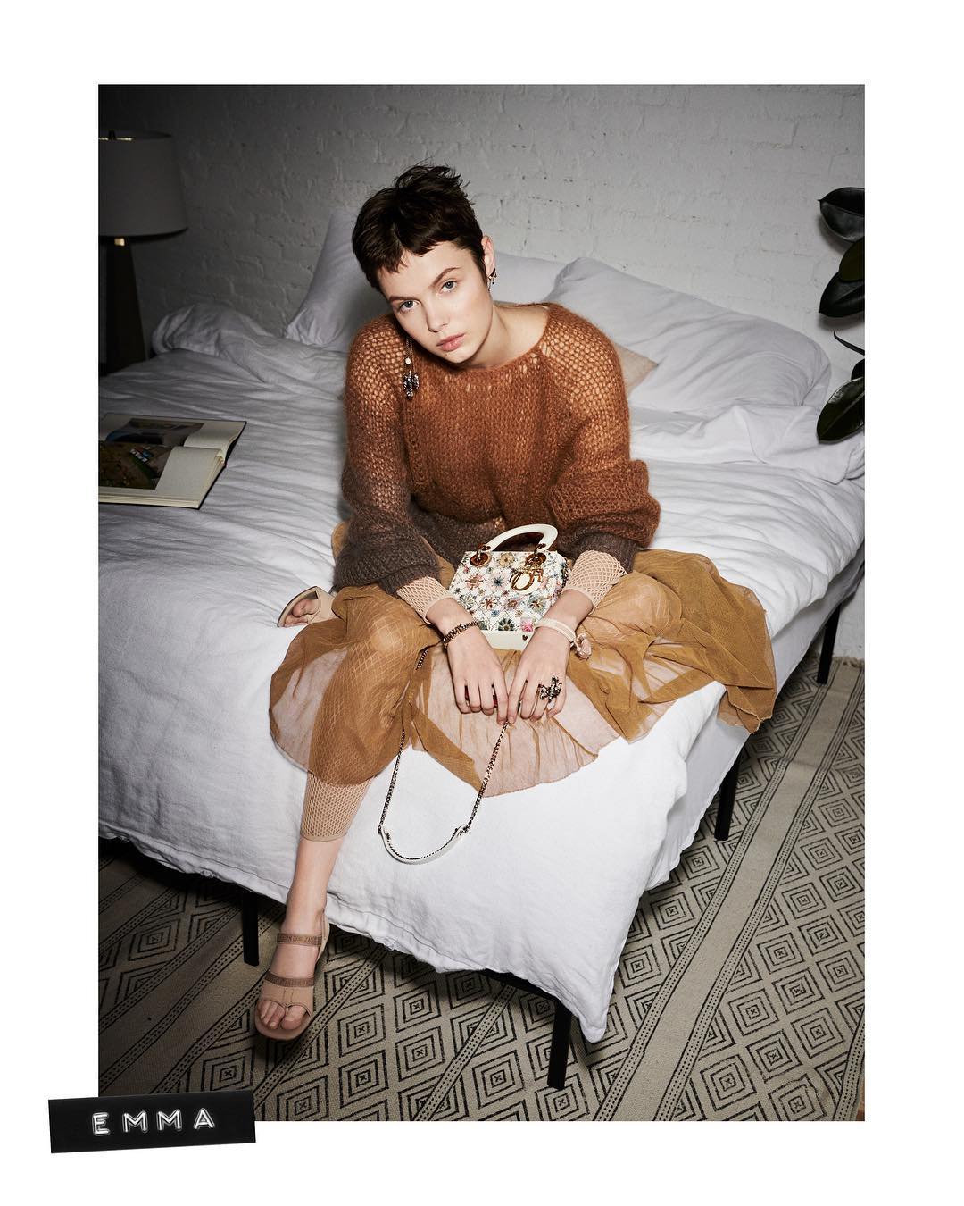 A fashionable woman with short brown hair sits on a white bed holding a decorative handbag. She wears a brown sweater and tulle skirt.