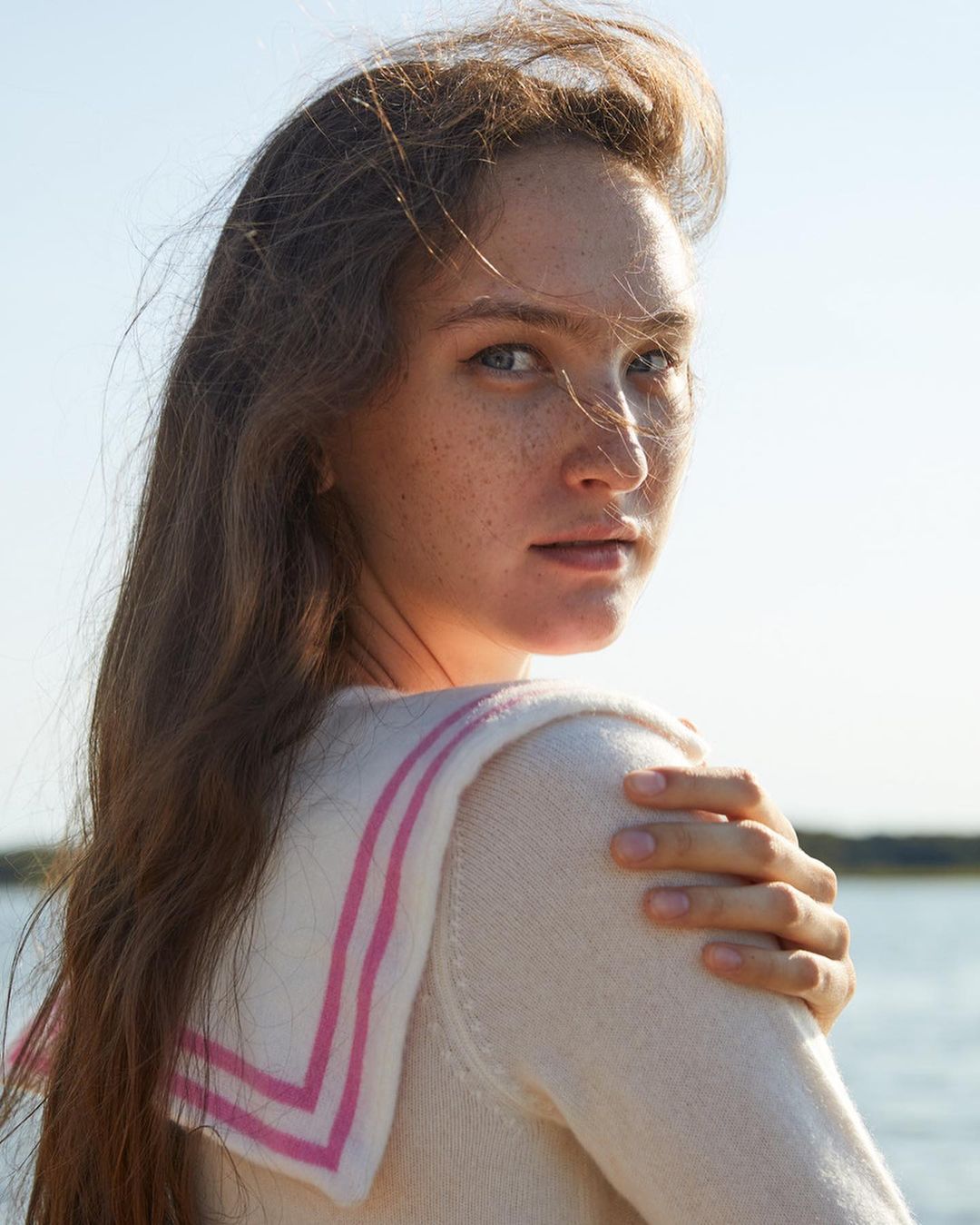 Portrait of a woman with long brown hair wearing a white sweater with pink stripes on the collar, standing in front of water.