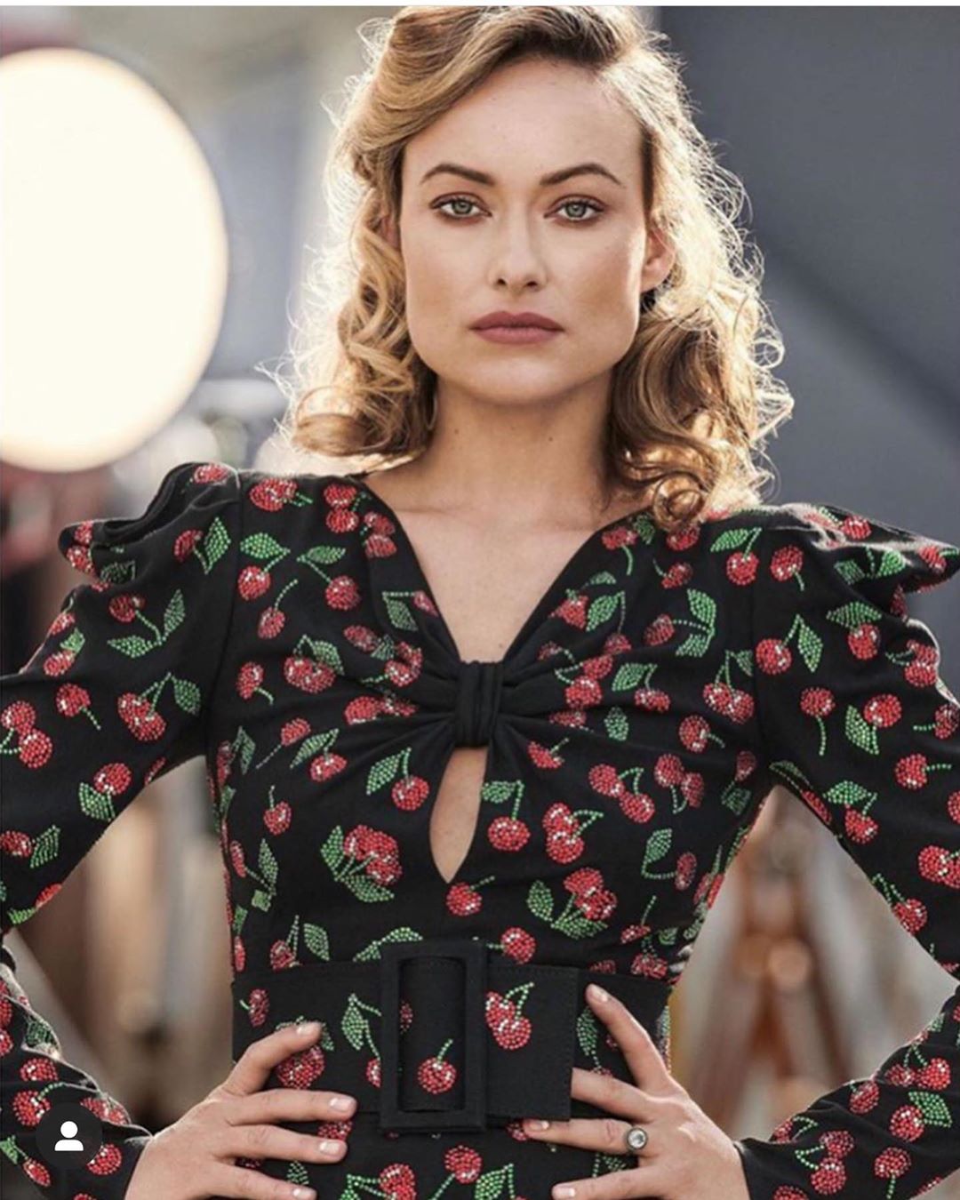 Close-up studio portrait of a fair-skinned woman in a black long-sleeve dress with red cherry print and curly blonde hair.