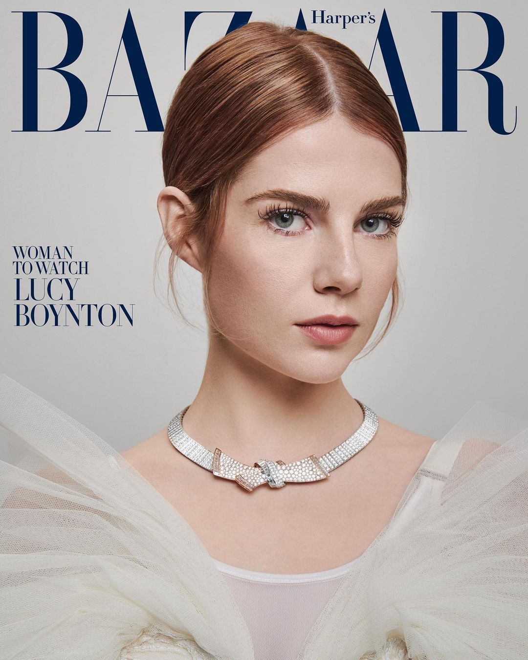 Close-up portrait of Lucy Boynton with red hair and a jeweled necklace against a grey background, as featured on Harper's Bazaar.