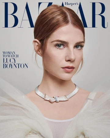 Close-up portrait of Lucy Boynton with red hair and a jeweled necklace against a grey background, as featured on Harper's Bazaar.
