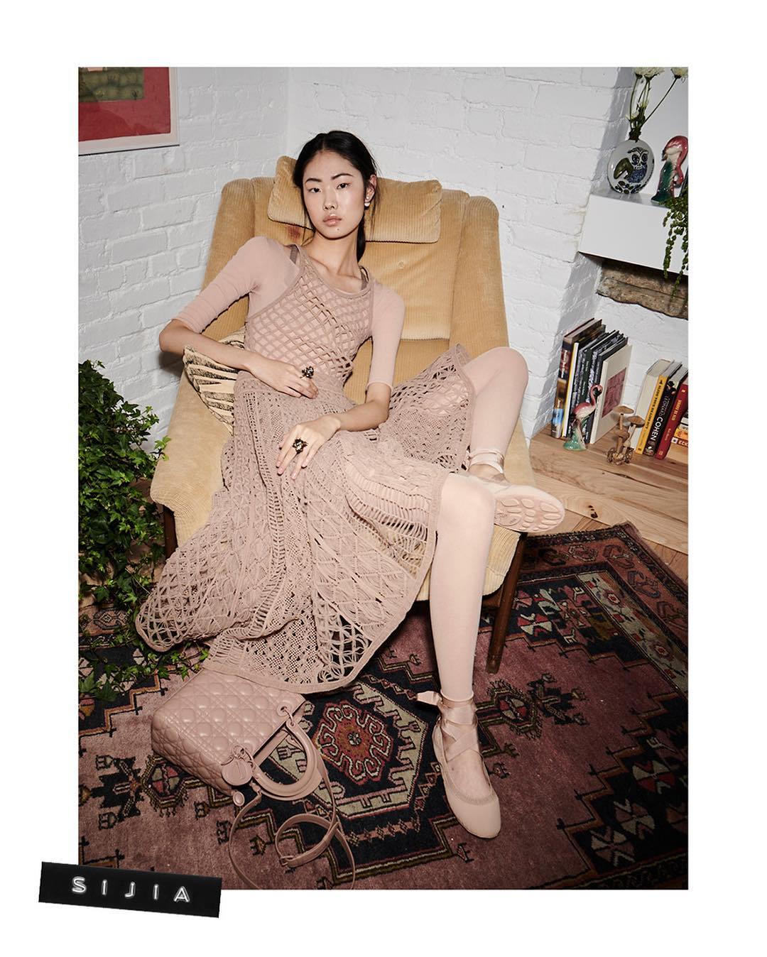 An Asian woman sits in a chair wearing a long, lacy dress, ballet slippers, and a purse. A patterned rug is on the floor.