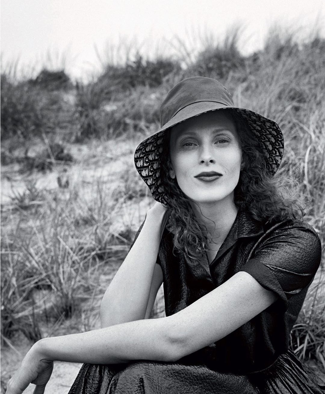 Monochrome portrait of a woman wearing a patterned bucket hat and dark collared dress outdoors against tall coastal grasses.