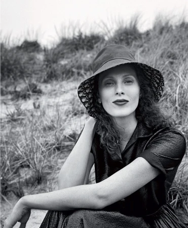 Monochrome portrait of a woman wearing a patterned bucket hat and dark collared dress outdoors against tall coastal grasses.