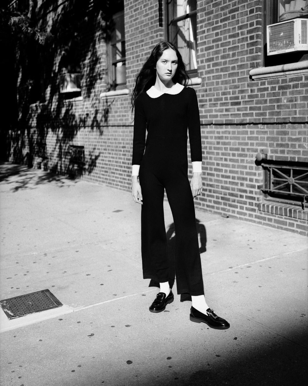 Woman in black jumpsuit and loafers stands on sidewalk in front of brick building, monochrome fashion editorial photography.