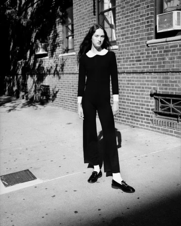 Woman in black jumpsuit and loafers stands on sidewalk in front of brick building, monochrome fashion editorial photography.