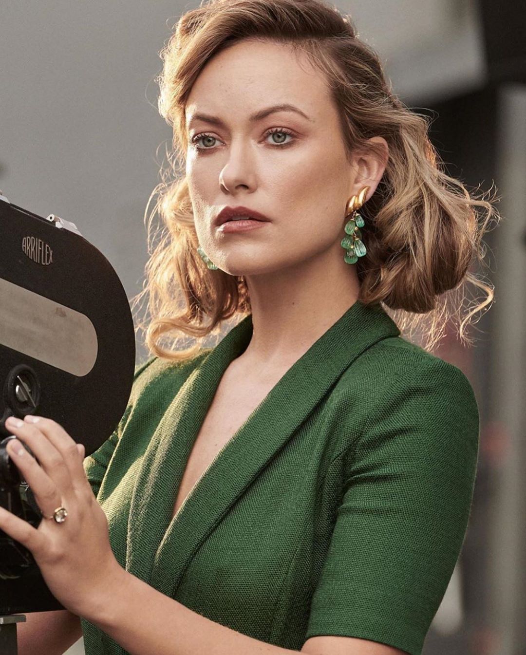 A woman with wavy blonde hair and green earrings in a green blazer next to a vintage camera. Studio lighting illuminates her face.
