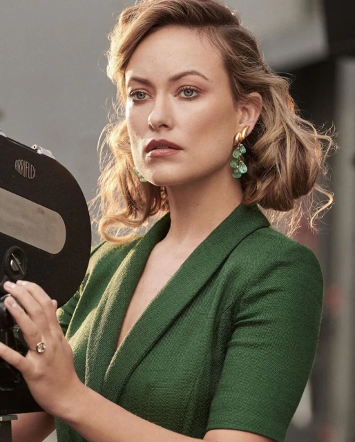 A woman with wavy blonde hair and green earrings in a green blazer next to a vintage camera. Studio lighting illuminates her face.