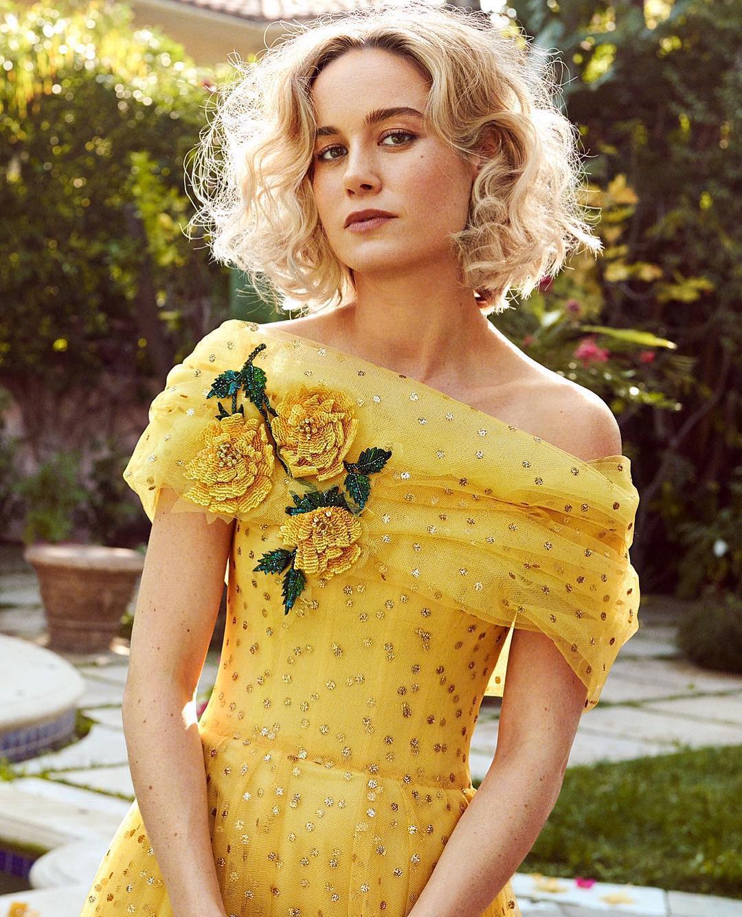 Brie Larson in yellow floral dress poses outdoors with curly blonde hair. Soft, natural light and greenery enhance the portrait.