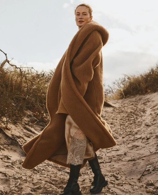 Adult female fashion model wears a long brown coat, dress, and black boots on a sandy beach with dune grass against a cloudy sky.