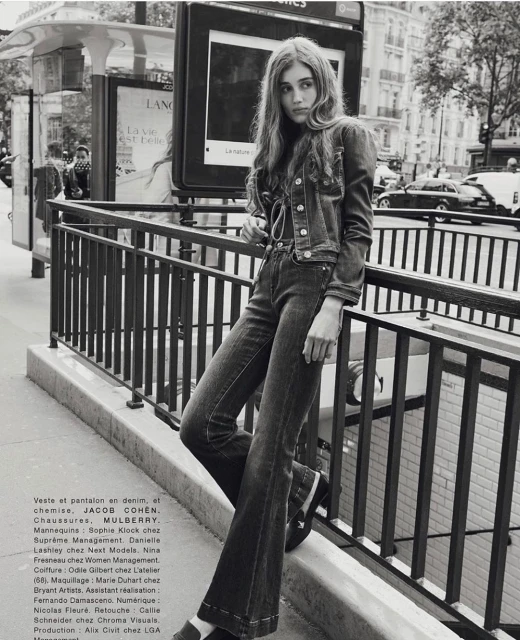 Outdoor fashion portrait in Paris of a young woman wearing a denim jacket and bell-bottom jeans leaning against a metal railing.