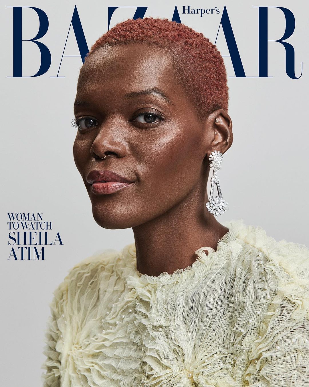 Close-up portrait of Sheila Atim on a magazine cover, featuring a ruffled cream-colored top and large chandelier earrings, against a solid gray background.