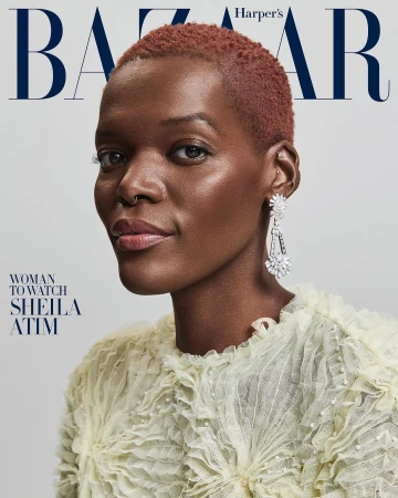 Close-up portrait of Sheila Atim on a magazine cover, featuring a ruffled cream-colored top and large chandelier earrings, against a solid gray background.