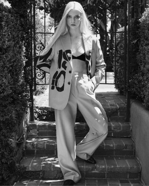Anya Taylor-Joy posing on brick steps in a beige suit with graffiti art, captured in a medium shot with an outdoor setting.