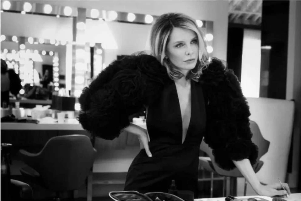 Black and white portrait of a woman in a black dress with large feathery sleeves, posing in a dressing room setting with mirror.