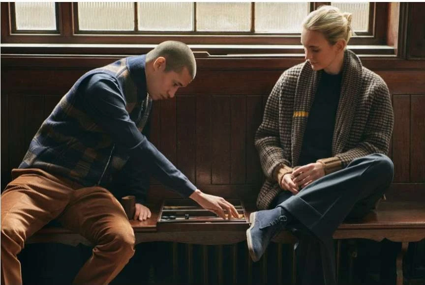 Two people playing backgammon indoors. A man in a vest leans over the game, while a woman in a patterned jacket looks on from a bench seat.