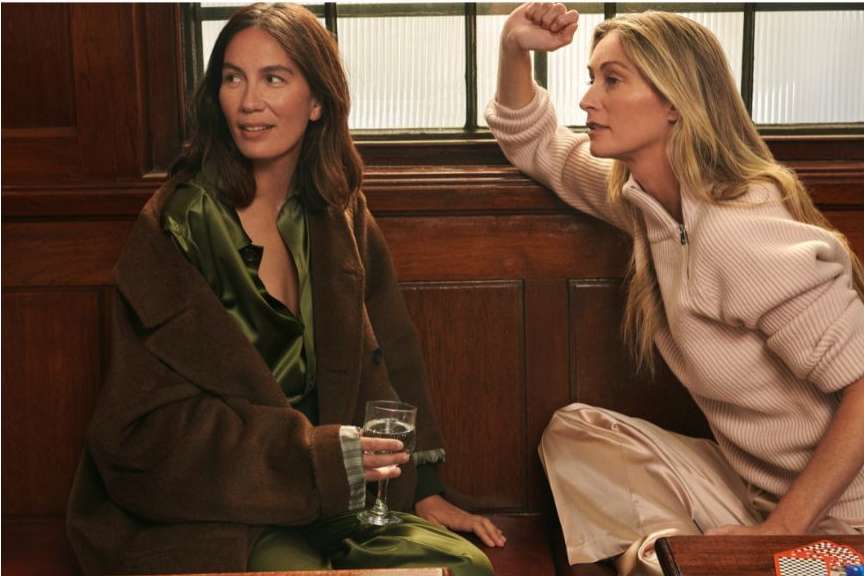 Two adult women in stylish casual fashion are seated indoors; one holds a wine glass, creating a relaxed, intimate atmosphere with warm lighting.