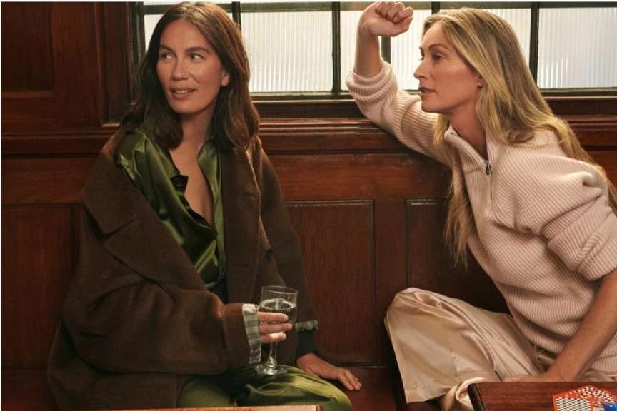 Two adult women in stylish casual fashion are seated indoors; one holds a wine glass, creating a relaxed, intimate atmosphere with warm lighting.
