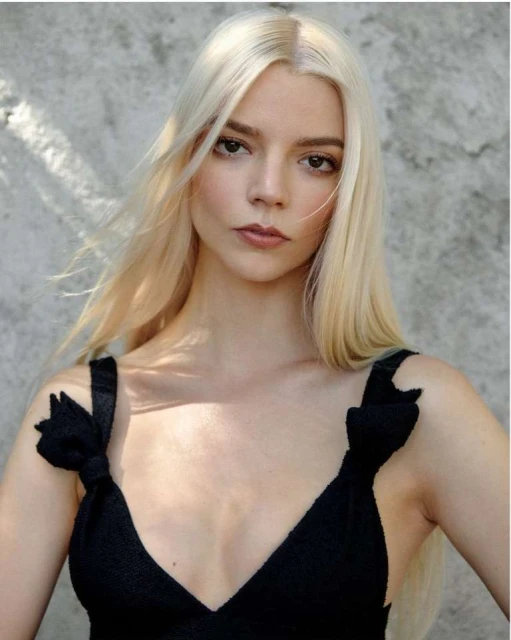 Close-up portrait of a woman with platinum blonde hair, wearing a black knit top, against a textured gray background in natural lighting.