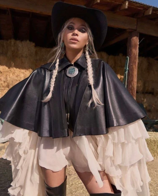 A woman in a black hat and cape-like jacket with a ruffled dress poses outdoors against a backdrop of hay bales and wooden structures.