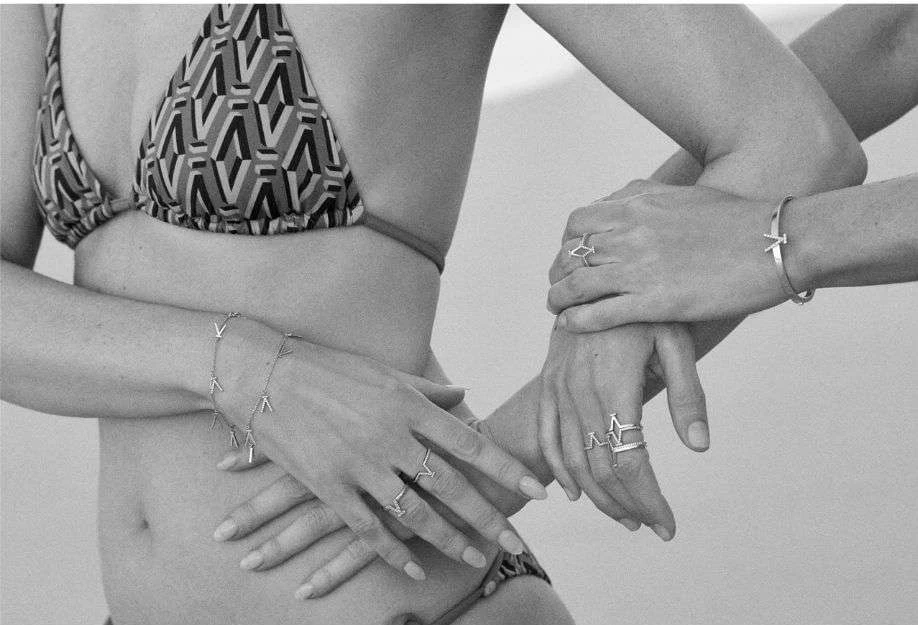 Two women pose in bikinis with hands stacked, showcasing rings and bracelets. The image is black and white with a shallow depth of field.