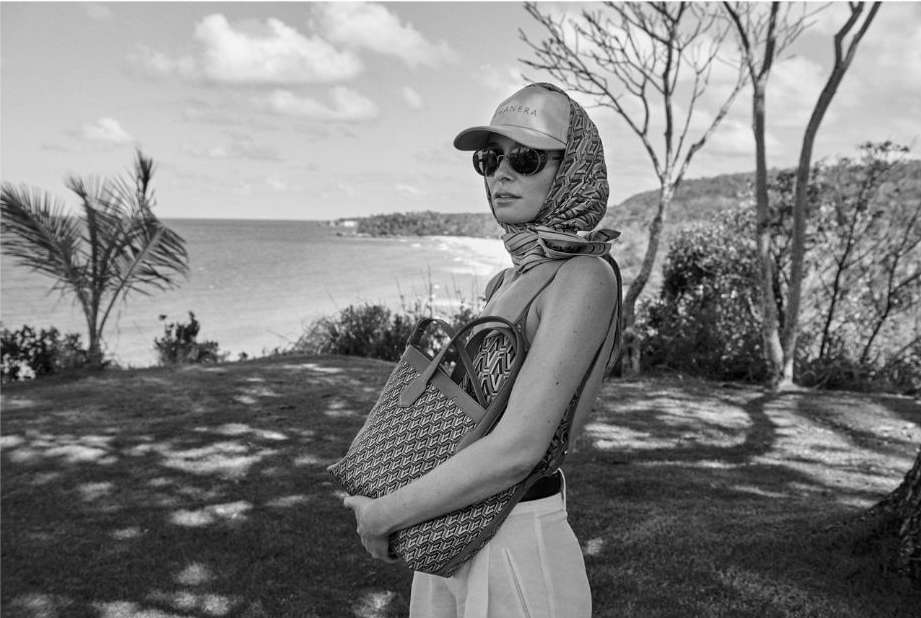 A woman on a grassy hill overlooking the ocean wears sunglasses, a patterned headscarf, and a baseball cap while holding a tote bag.