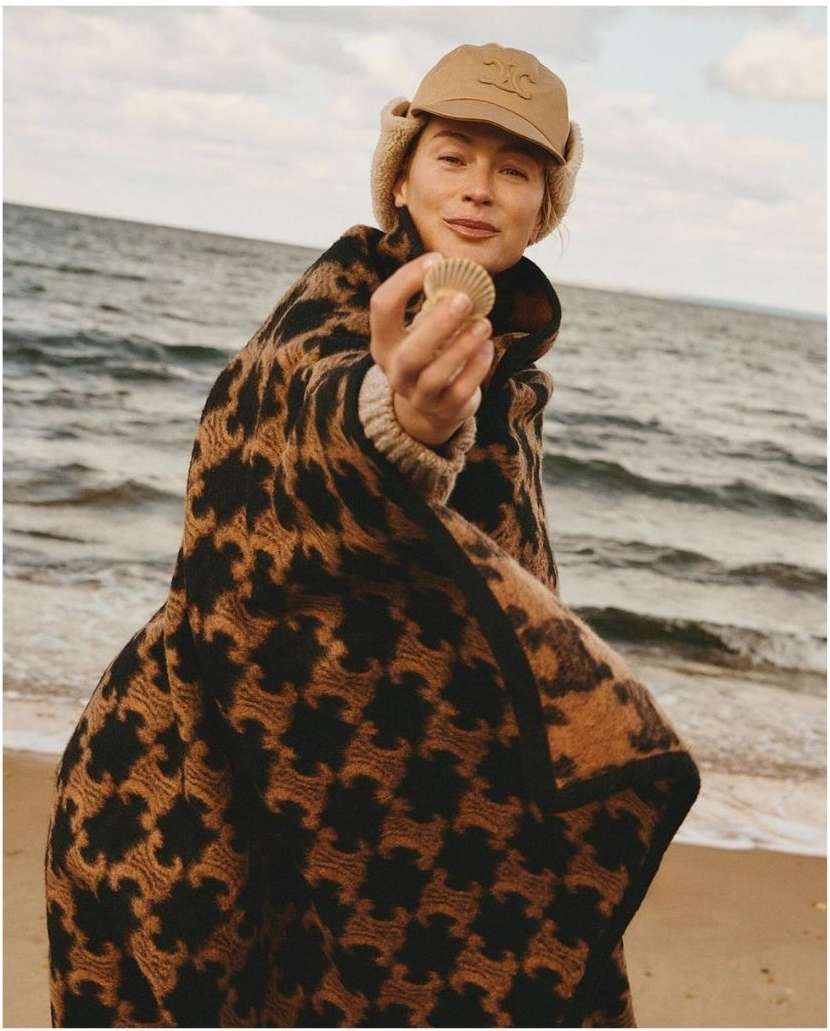 A woman on a beach holds a shell up to the camera, wearing a patterned brown poncho, beige cap, and ear muffs, with the ocean behind her.