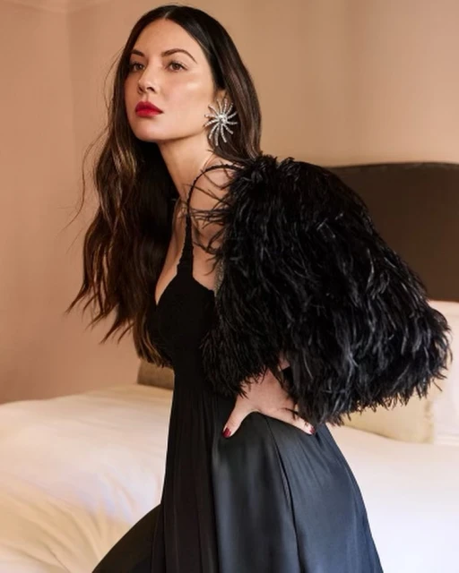 Elegant woman in a black dress with a feather accessory posing indoors. The image features a formal portrait against a neutral backdrop.