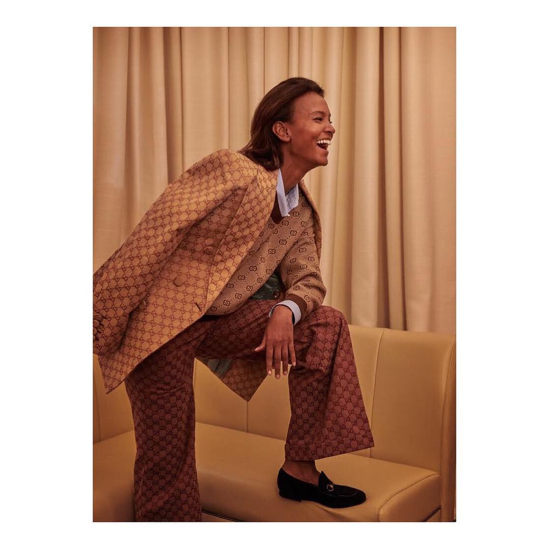 A woman with short hair laughs, wearing a patterned brown blazer and pants, standing on a yellow sofa in front of tan curtains.