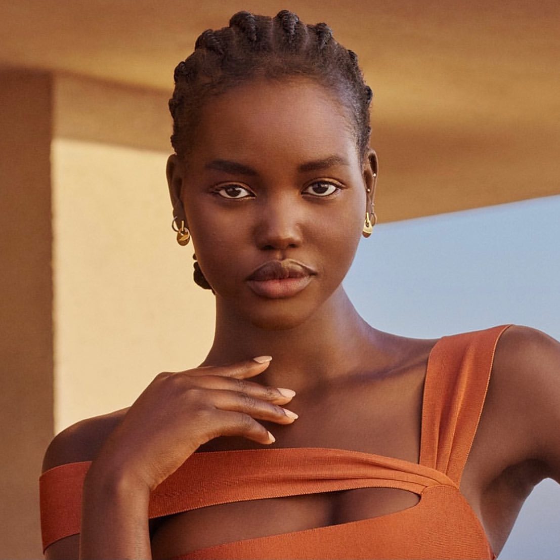 Close-up portrait of a dark-skinned woman with braided hair wearing an orange, modern-cut top against a neutral color gradient backdrop.