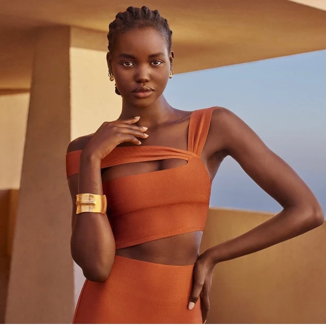 Elegant dark-skinned woman with braided hair posing in an orange cut-out dress with gold bracelet against a blurred architectural background.