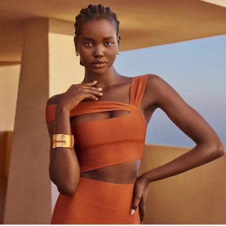 Elegant dark-skinned woman with braided hair posing in an orange cut-out dress with gold bracelet against a blurred architectural background.